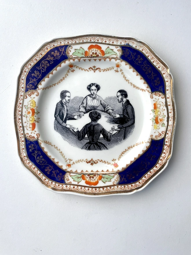 a vintage plate with ornate border has the vintage illustration of 2 men and 2 woman holding a seance at a table