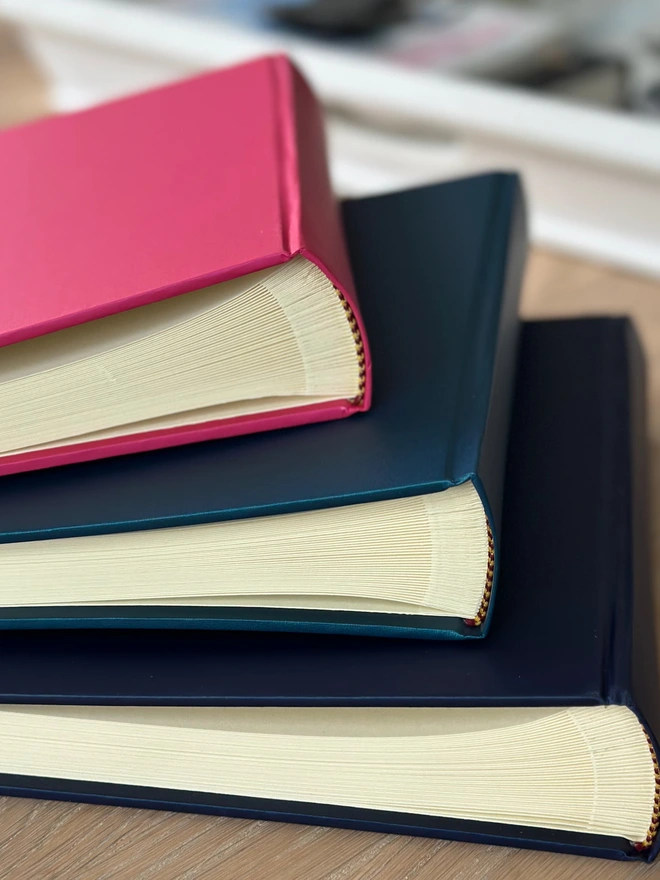 A pile of hardback photo albums rests on a wooden surface. The colours are pink, blue and black.