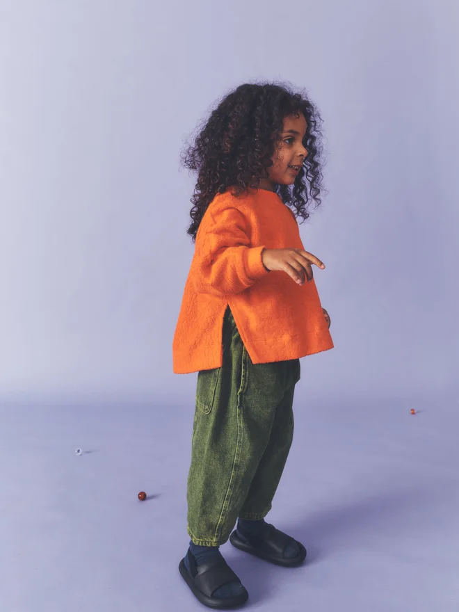 Side view of a child against a lilac backdrop, wearing a Studio Koter orange textured fleece top with side splits and green acid-wash barrel-leg jeans.