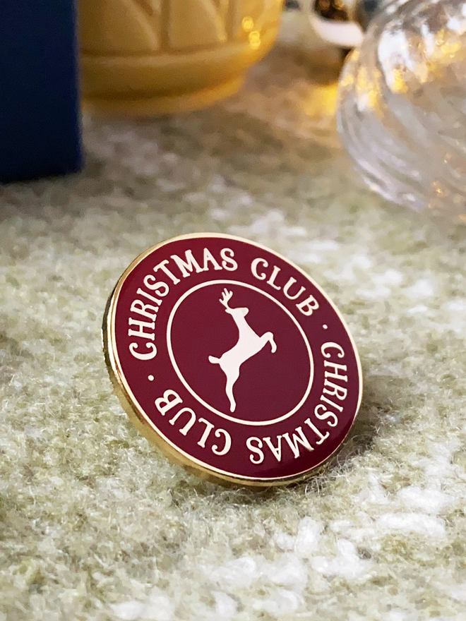 A deep red and gold enamel pin badge with a gold reindeer in the centre and the words "Christmas Club" around the outside is on a green blanket.