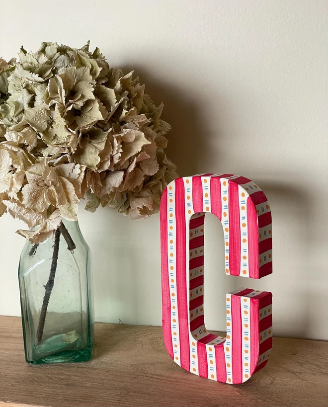 A hand-painted decorative initial letter  with stripes on a wooden table against a light wall. 