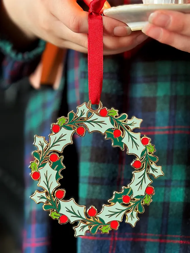 A child wearing a tartan dress holds a vintage-style enamel Christmas wreath ornament with green holly leaves and red berries, hanging from a red ribbon, alongside a small white candleholder with a lit candle.