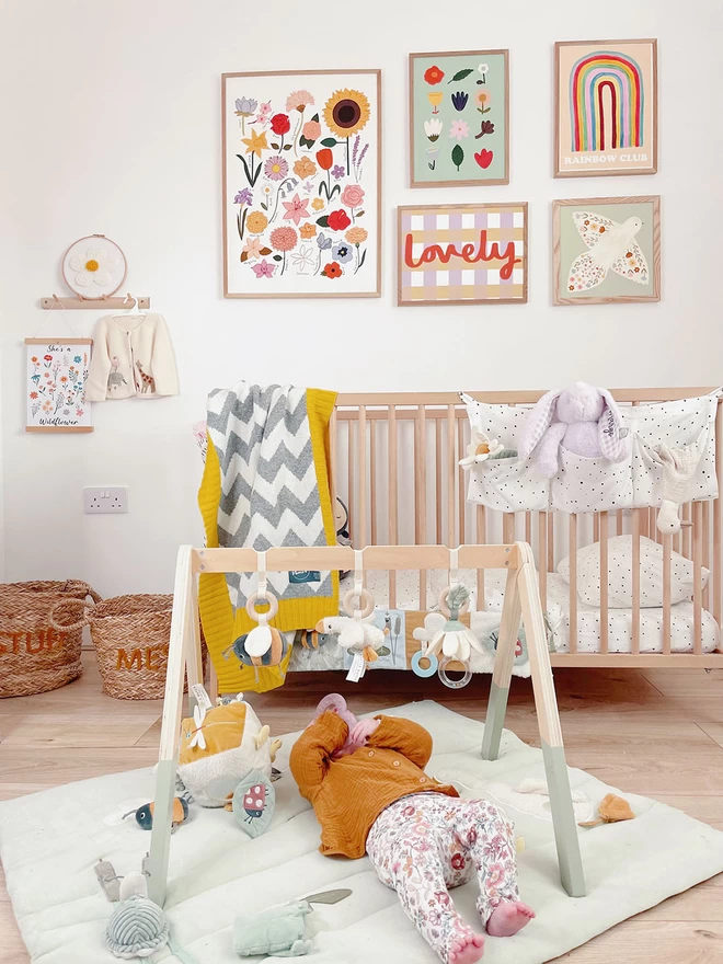 A baby lies on its back on a playmat beneath a baby gym in a bright and colourful nursery. In the background is a wooden cot with a grey and white zigzag baby blanket draped over the side. Modern prints line the walls and toys are scattered on the floor.