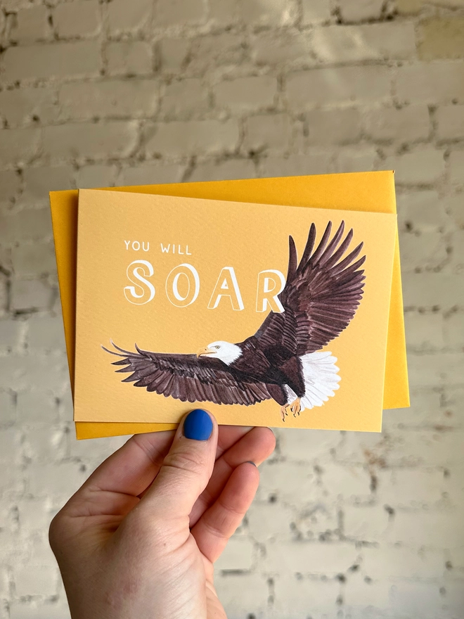 a bright yellow greetings card featuring an illustration of a bald eagle with the text “you will soar” with a deep yellow envelope