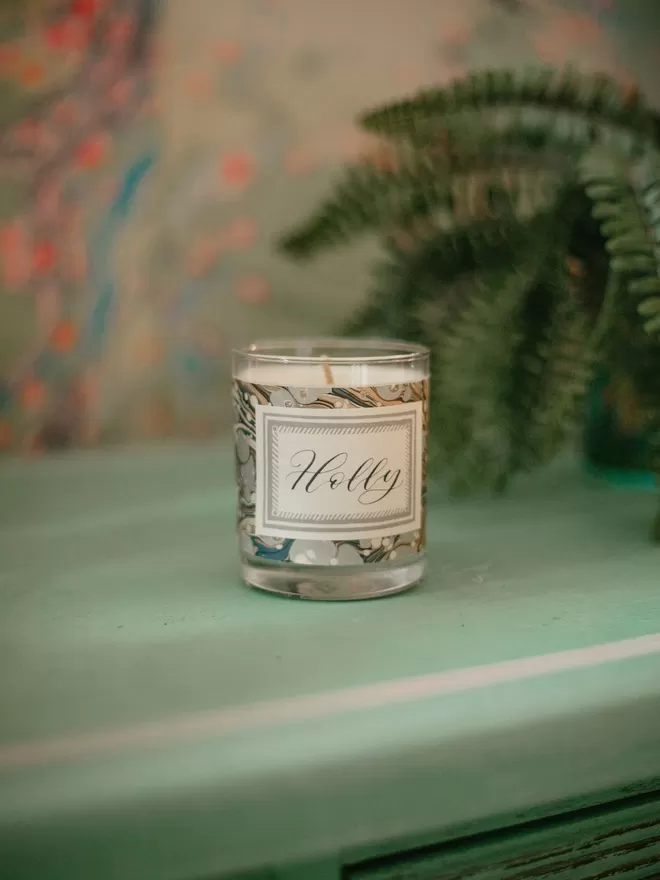 Sassigraphy Hand Poured Scented Candle With Personalised Marbled Label seen on a green countertop.
