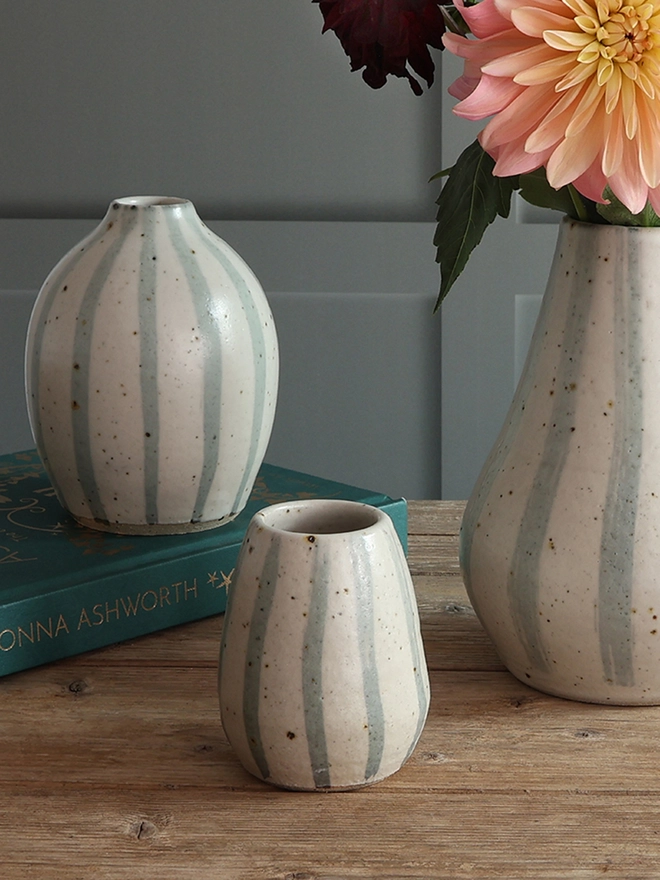 Handmade Ceramic Stoneware Vases in three sizes: small, medium and large. The vases sit on a wooden surface alongside a book and pink sea urchins. The largest vase is filled with a large bouquet of flowers. 