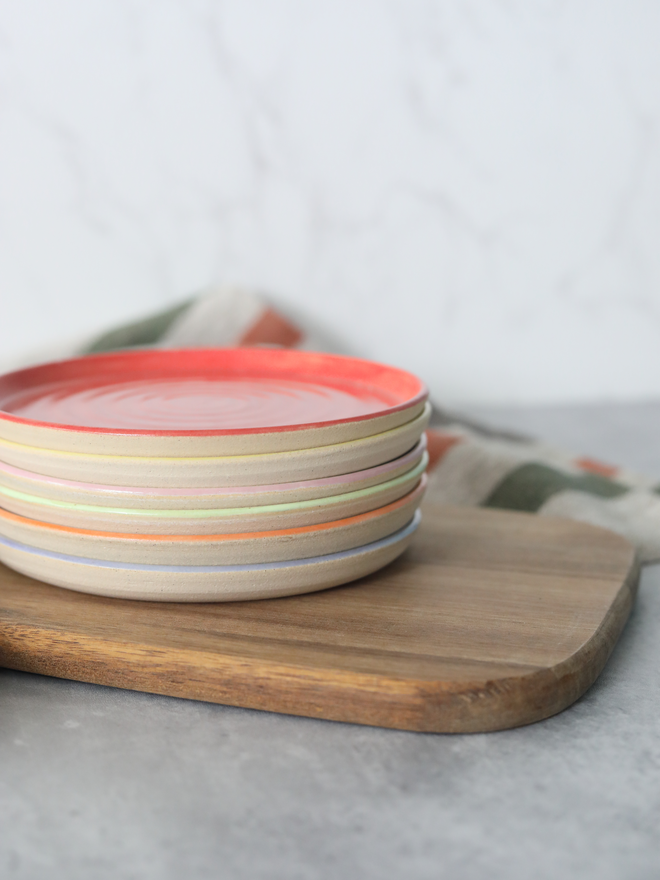 A close-up stack of handmade ceramic cake plates on a wooden board, topped with a cherry red plate, with multicoloured rim edges visible beneath in yellow, blush pink, spring green, mandarin orange, and lavender, styled with a linen cloth against a white marble background