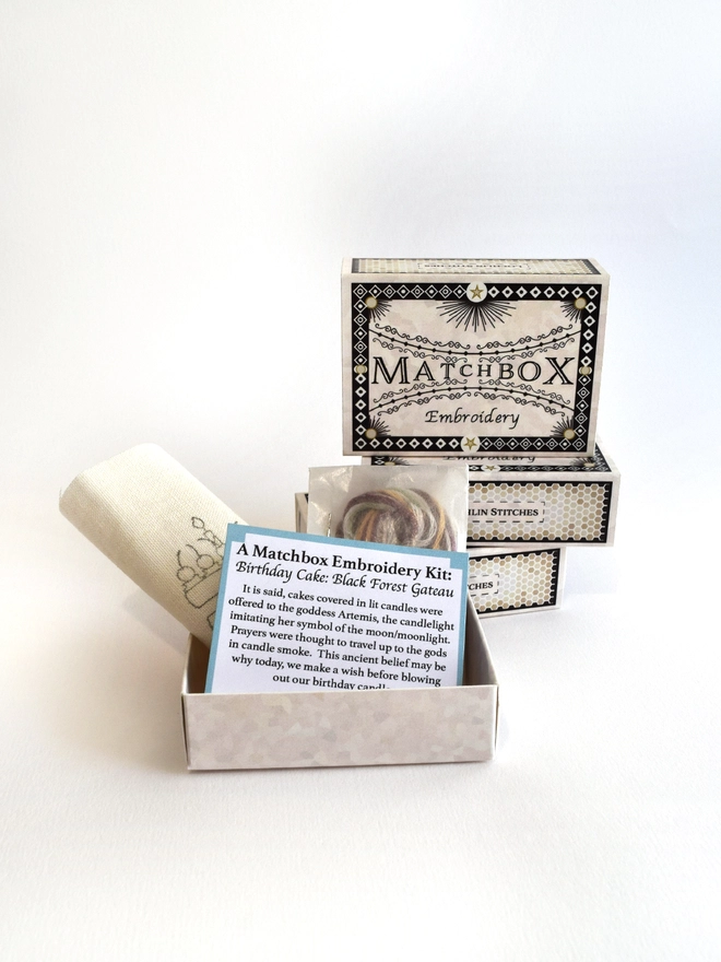 The kit supplied in a beautifully designed matchbox, pre-printed fabric and small instruction leaflet neatly folded to sit inside, along with the yarns and needle housed in a small glassine packet.  A stack of matchboxes sit in the background.