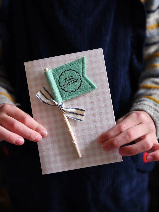 A child is holding a Personalised Father’s Day card with green felt flag reading “Elsie & Daddy”, handmade keepsake card with ribbon and wooden stick detail.