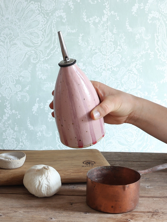 A female hand holds a Handmade Ceramic Stoneware Oil Pourer in Pink Stripe above a wooden surface. The oil pourer bottle is surrounded by bulb of garlic, a brass measuring cup, a wooden chopping board and small ceramic dishes containing salt and pepper. 