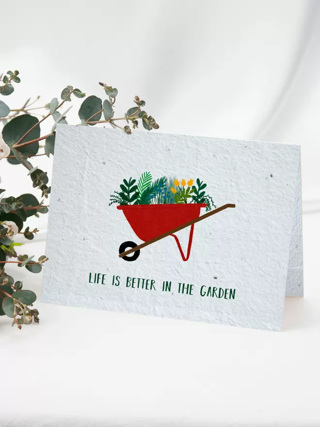 Seeded Paper Greeting Card featuring an illustration of a wheelbarrow full of flowers and plants with ‘Life is better in the Garden’ written underneath with a bunch of flowers in the background