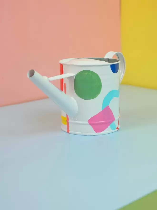 Hand painted watering can by Julie-Anne Pugh. Base colour is white with bring coloured shapes weaving across the body of the can on a yellow and orange background. Colours on shapes include red, violet, peach, light blue, royal blue and green.