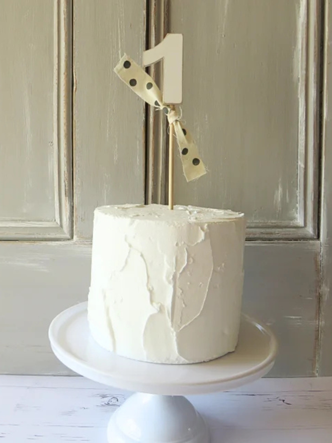 hand painted wooden number cake topper. a white number one cake topper with a spotty ribbon on a stack of donuts.