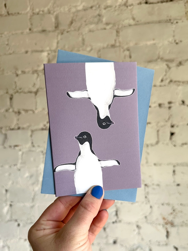 a purple greetings card featuring two illustrations of a penguin with a blue envelope