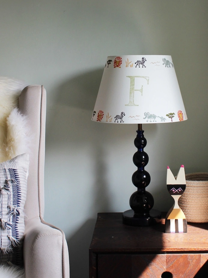 A cosy corner of a child's bedroom with a wooden nightstand, a blue lamp with a safari-themed shade, and a wooden cat figurine. Beside it, a cream armchair with a fluffy throw and textured cushion sits under a framed botanical print. A nearby shelf holds colourful toys, while sunlight casts warm shadows.