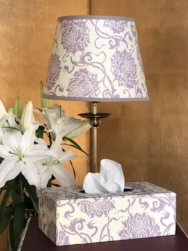 Retro Drum Lampshade. A lilac and cream floral block-printed lampshade on a brass lamp with a matching patterned tissue box and white lilies. The coordinating hand-printed pieces create a soft, elegant look for a bedroom or dressing table.