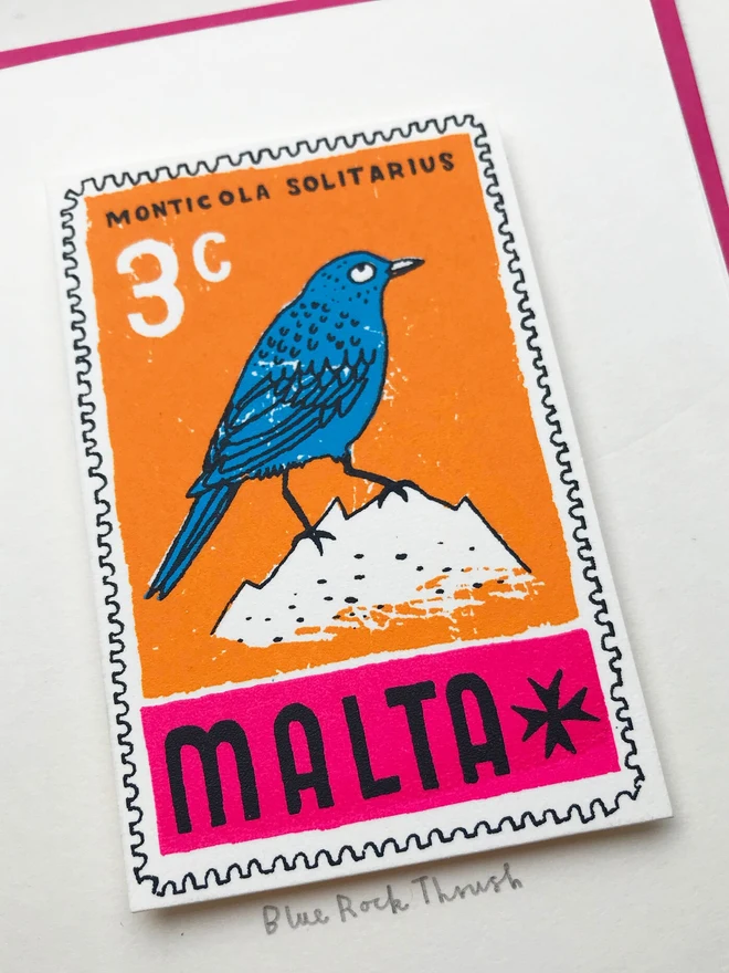 Bird Stamp Card Malta/ Blue Rock Thrush, a white card with a colourful bird design in the centre on top of a pink envelope on a white backdrop. 