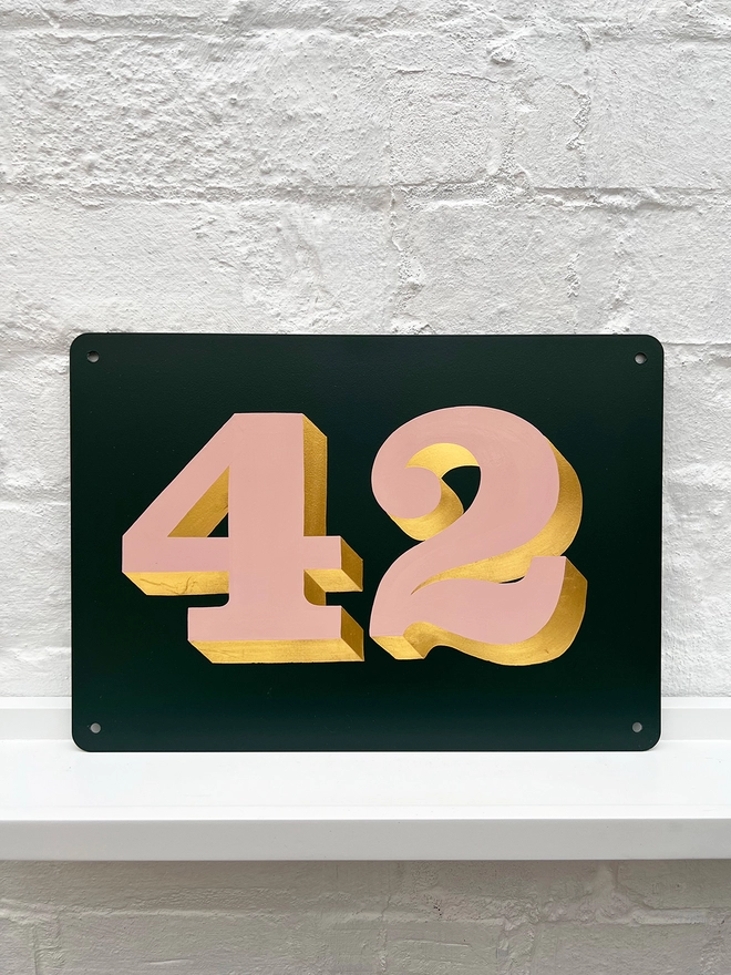 Hand painted house number against a white brick wall, number 42 in serif style in dusky pink and directionally burnished 23 carat gold leaf, on anthracite grey background. 