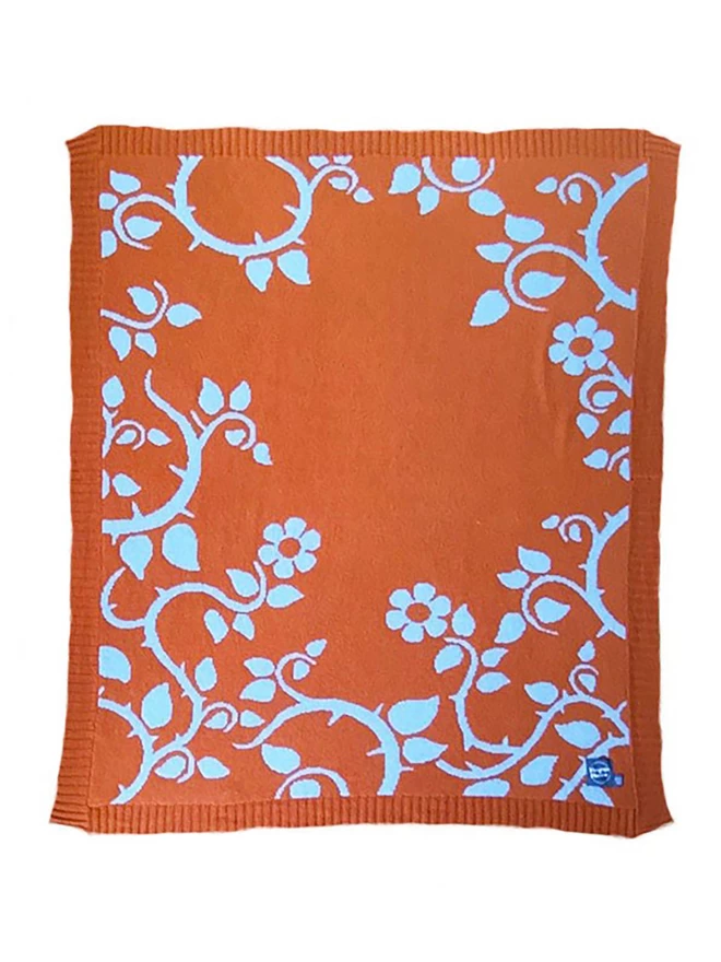 Product shot showing the front of the rust briar rose junior blanket, rust orange background with muted blue trailing flower  and thorn pattern.