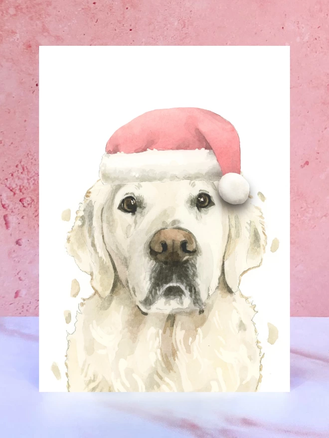 A Christmas card featuring a hand painted design of a cream golden retriever, stood upright on a marble surface surrounded by pompoms. 