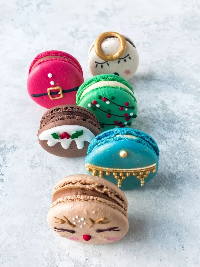 several decorated macarons themed like Christmas character - fairy lights, rudolf the red nosed reindeer, Christmas bauble, angel, Santa and Christmas pudding are arranged  on a white background