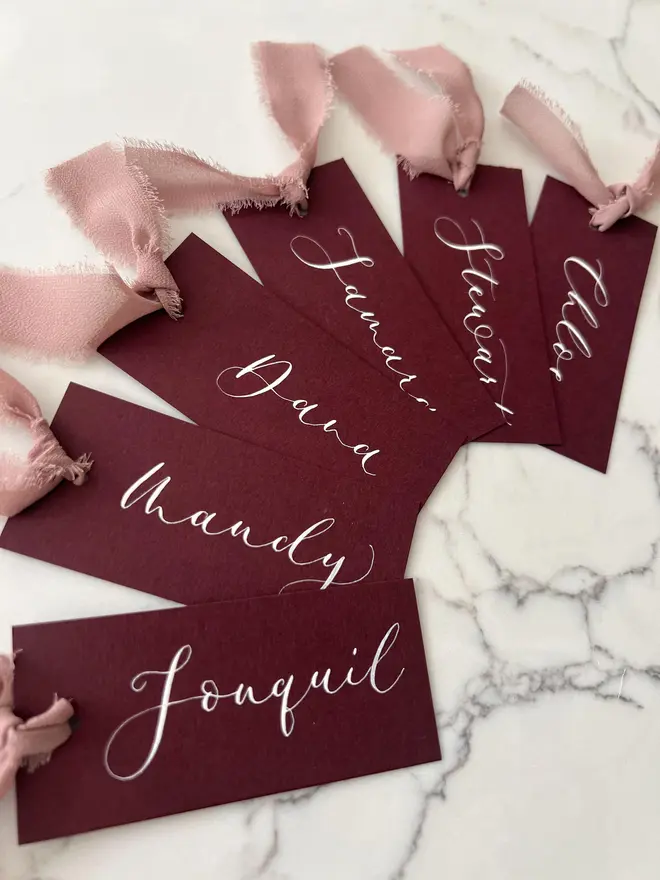 Personalised Calligraphy Wedding Burgundy Place Cards, burgundy place cards set together on a wooden table with chiffon plum ribbons. 