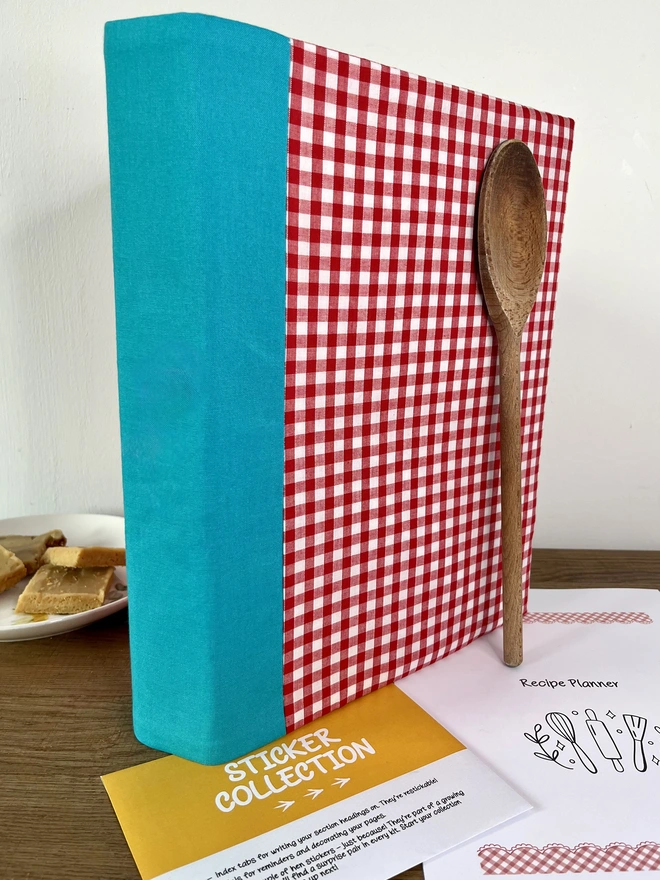 Red gingham recipe binder with plain (non-embroidered) teal spine, standing on kitchen table with wooden spoon propped against it and a plate of baking beside the folder.