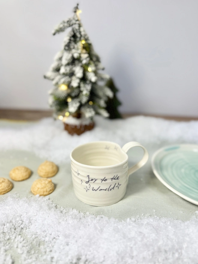 cream hand thrown porcelain mug with the words 'Joy to the World' printed onto the side with pretty line drawn stars. Sitting on a tabletop with snow, a mini lit Christmas tree and some biscuits