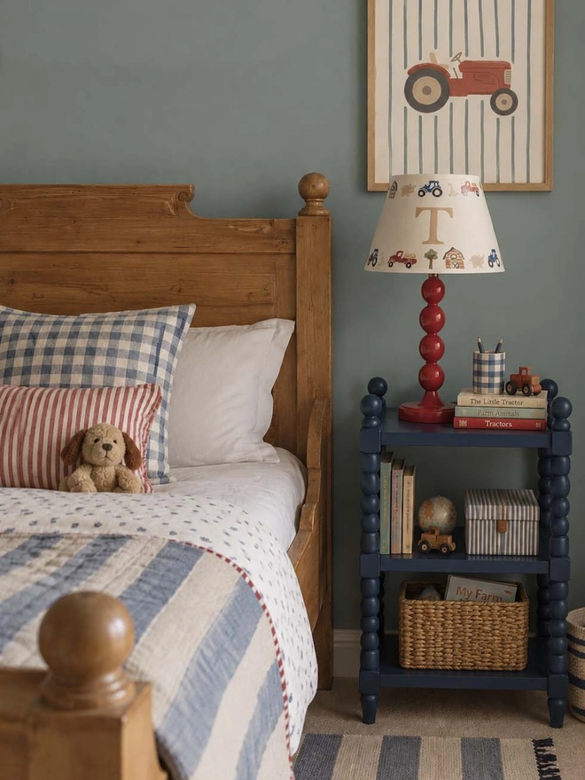 Personalised farmyard lampshade with initial T on a glossy burgundy red bobbin table lamp, styled on a navy blue bedside table in a cosy boy's bedroom. The hand-printed cream shade features tractors, farm animals and barns, alongside a wooden bed, blue gingham bedding, striped cushions and vintage-inspired tractor artwork.