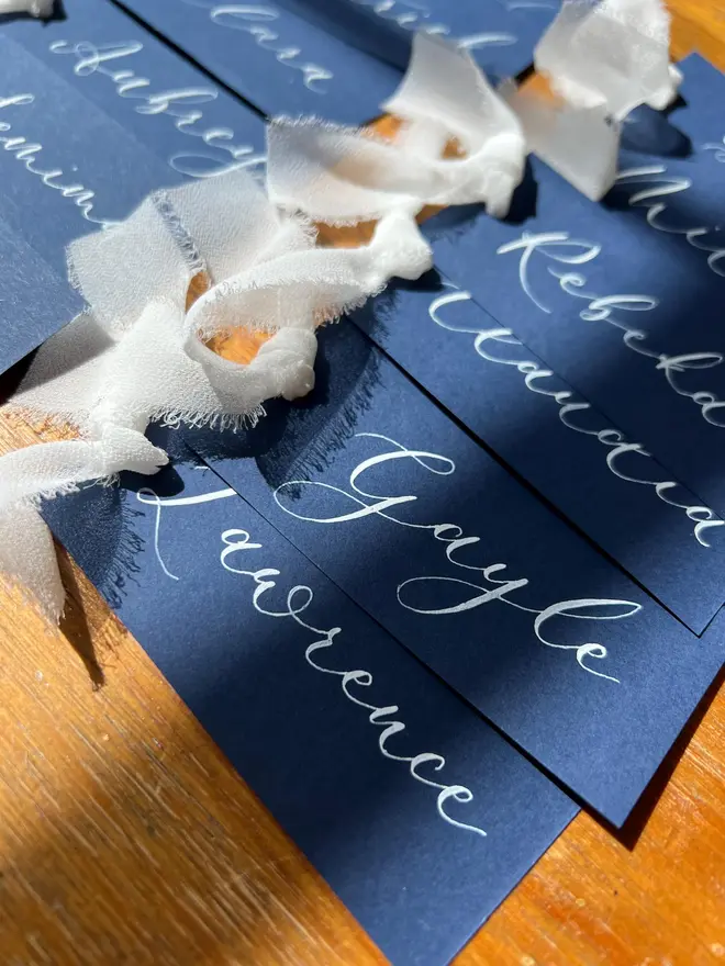 Personalised Calligraphy Wedding Navy Place Cards, navy place cards set together on a wooden table with white ribbons. 