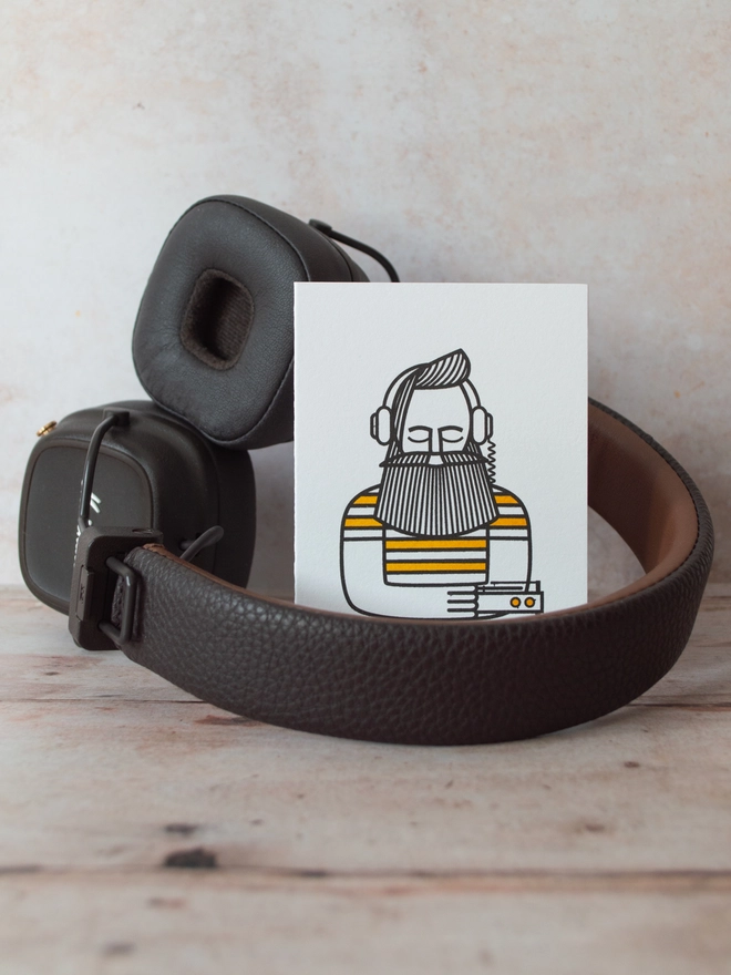 a modern illustration of a man, eyes closed listening to music through headphones is printed onto a small greeting card which sits next to some headphones.