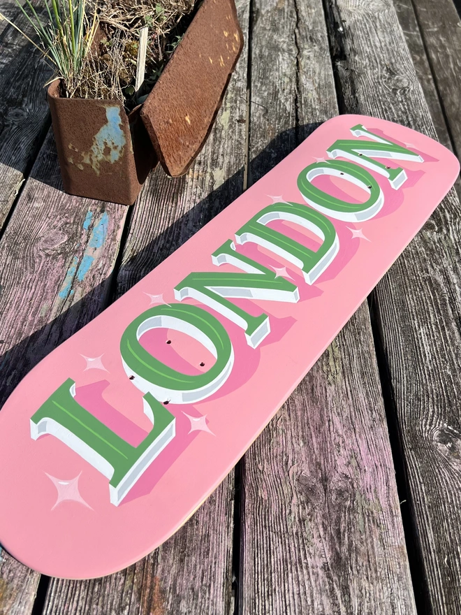 Pink skateboard deck with 'LONDON' in sage green lettering, white and grey shade and light pink sparkle details.