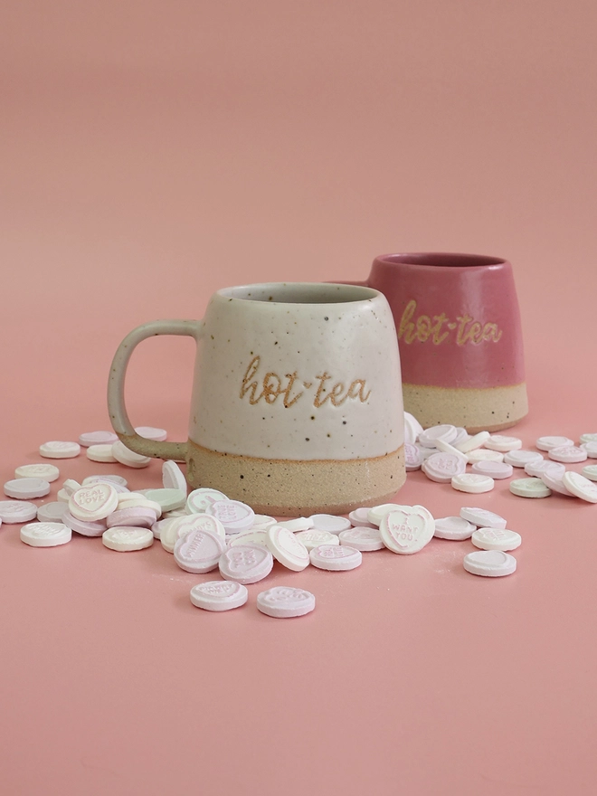 two handmade mugs which feature the words hot tea sit on a pink backdrop surrounded by lots of love heart sweets 