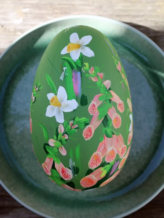 Soft green floral hand painted wooden egg, adorned in foxglove and daffodils positioned on a green plate