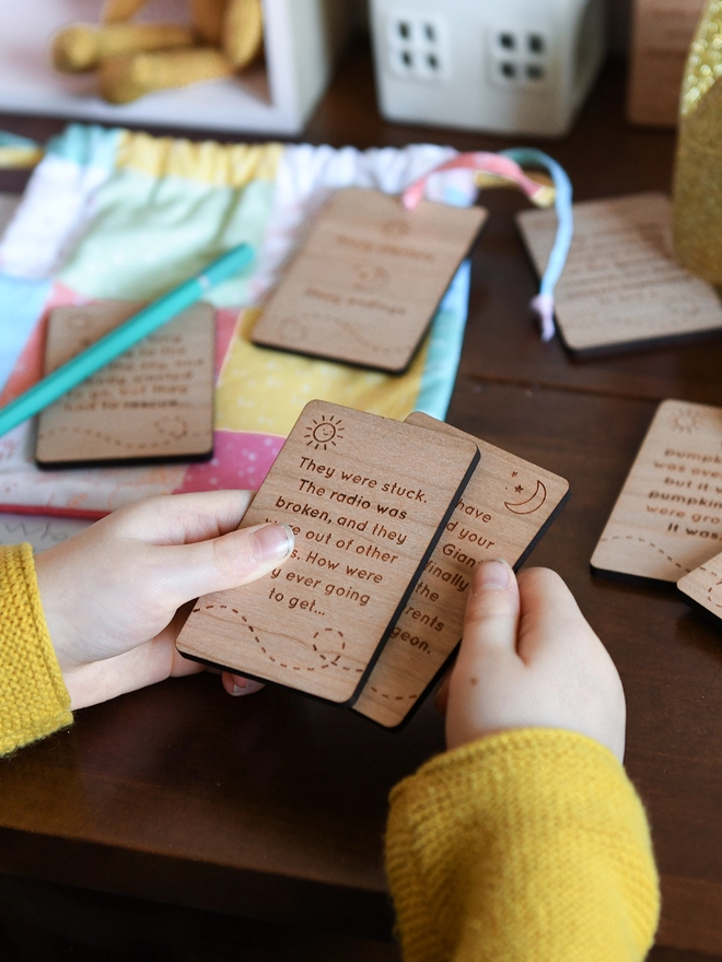 A child's hands hold two wooden story cards above a wooden desk. Each card has a story idea engraved on it, other cards lay on the desk behind.