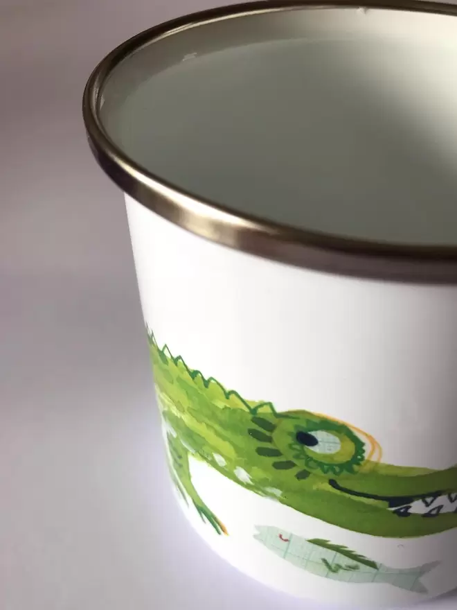 Close up of a white enamel mug with a green crocodile illustrated design