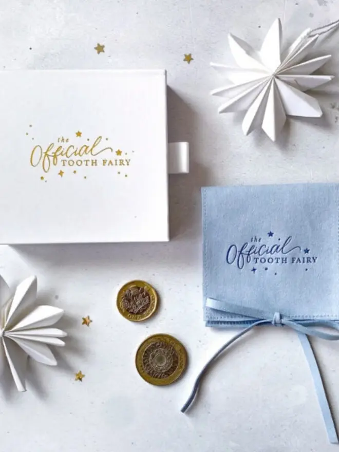 A white gift box with the gold foil logo lies beside a pale blue suede pouch tied with a bow. Two pound coins and gold confetti stars are scattered between them.