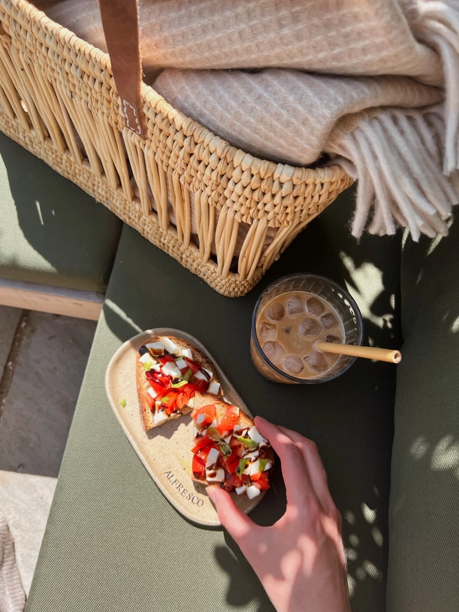 hand holding a piece of bruschetta above an oval ceramic plate with the word 'alfresco' stamped around the edge