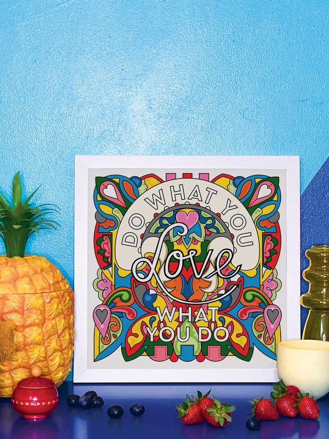 “Do what you love, love what you do” is written over a vibrant, rainbow coloured illustration, in a white frame propped against a wall painted light and dark blue, on a shiny blue cabinet. Next to the frame is a plastic pineapple ice bucket, a small wooden red pot a handful of blueberries, a yellow glass vase, a small yellow ceramic pot and 4 strawberries.