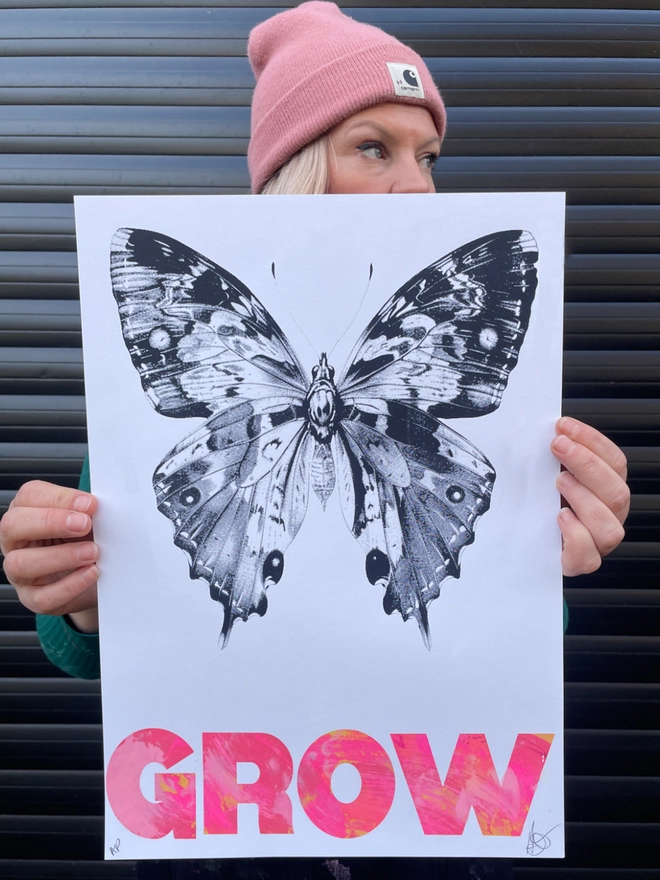 Woman artist with pink beanie holding an original artwork of a Butterfly print, with the word grow mono printed