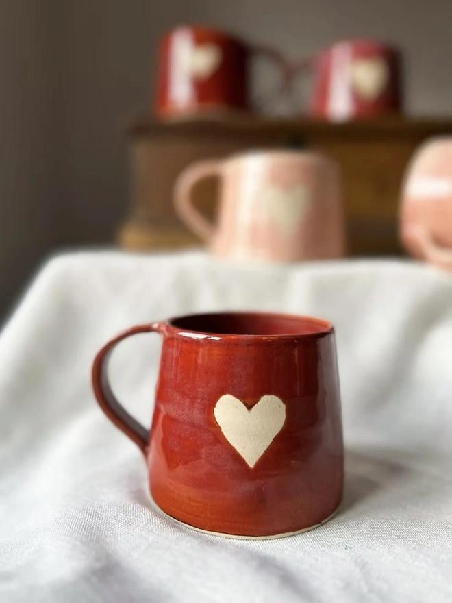 Heart Mug Cinnamon, a heart mug on a white surface in front of a group of more pink and red heart mugs. 
