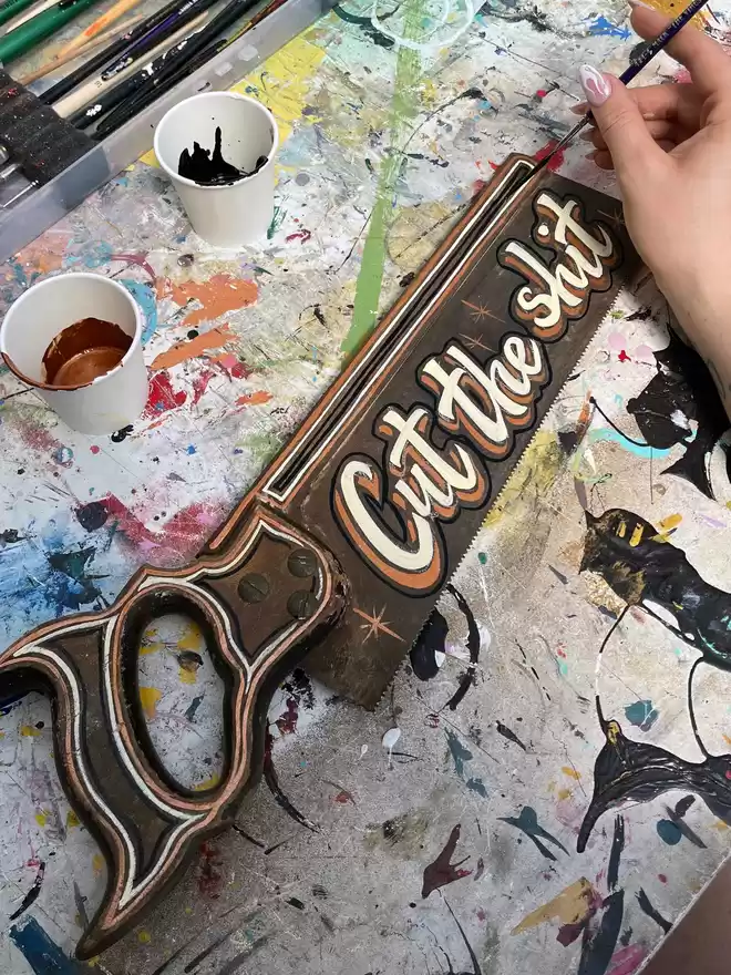 Hand-painted vintage saw spelling 'Cut the shit' in ivory and bronze with black details.