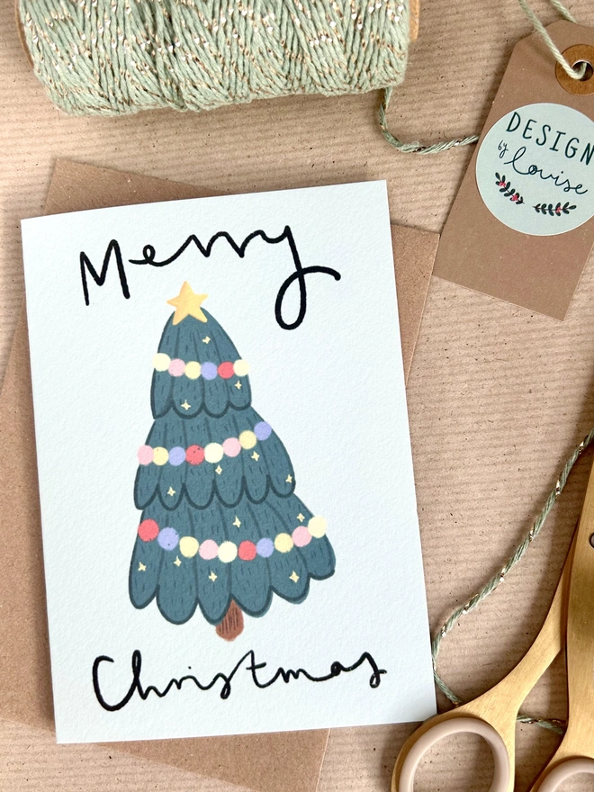 A green Christmas tree decorated with pastel coloured pompom tinsel.  Printed on a pale blue background. The words Merry Christmas are written in cursive handwriting around the tree.