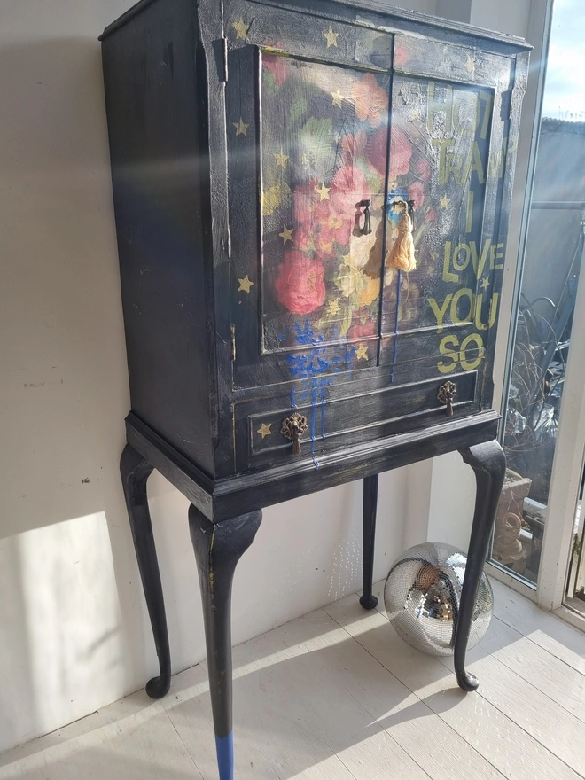 Elegant cocktail cabinet in black with colourful florals and text in chartreuse green that reads 'hot tramp, I love you so', a David Bowie lyric. Heavily distressed to age it. The interior is cobalt blue.