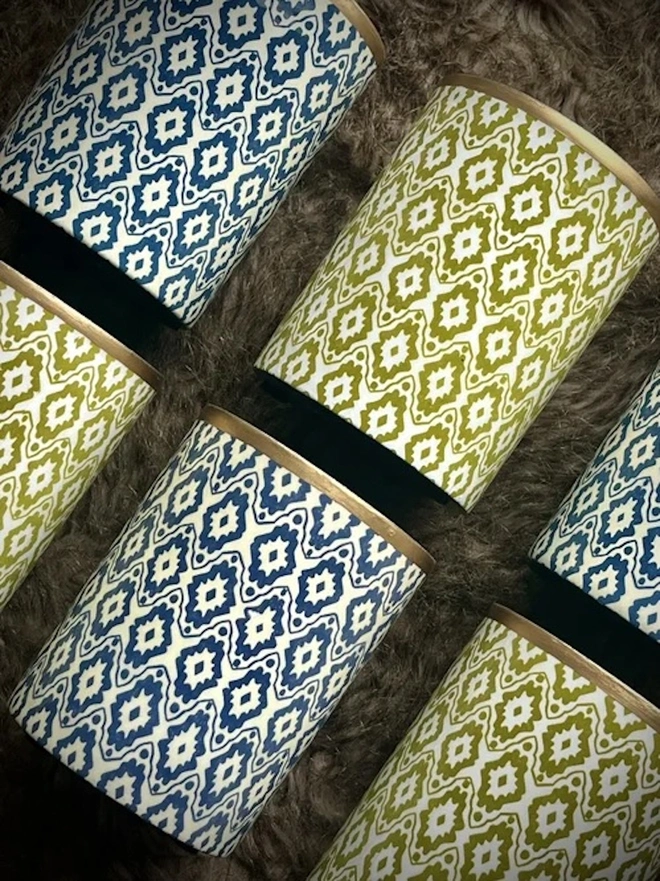 Perfect Enamel Pot. A collection of green and blue ikat-patterned pots with gold rims laid on a soft brown faux fur throw. The matching geometric designs show the range of colourways available for decorative storage.