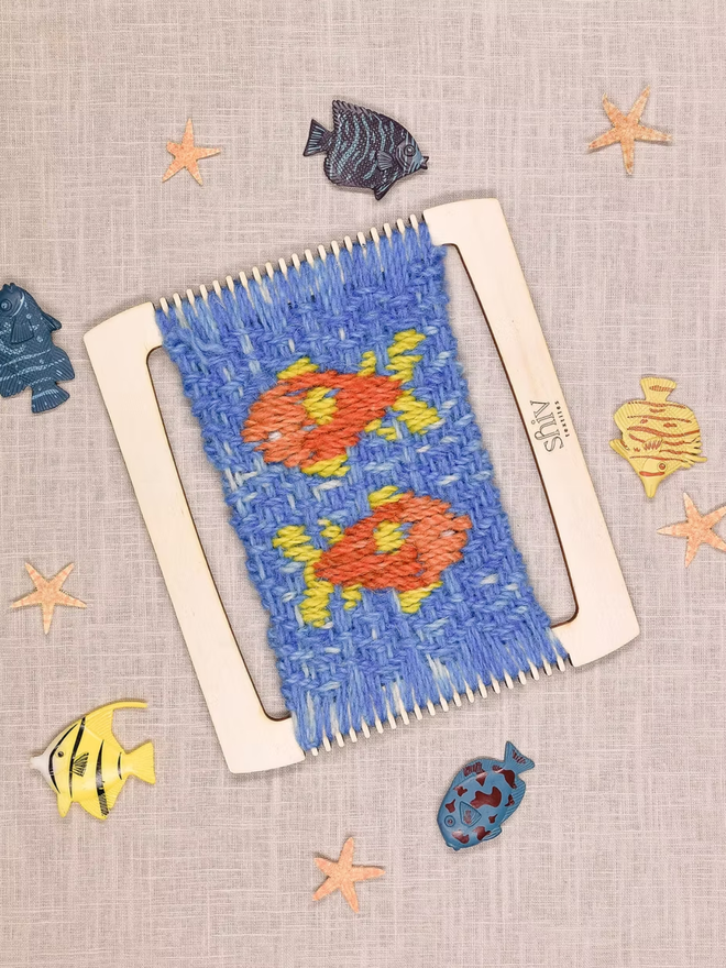 seaside weaving kit. a blue weave piece with orange fish photographed on a loom.