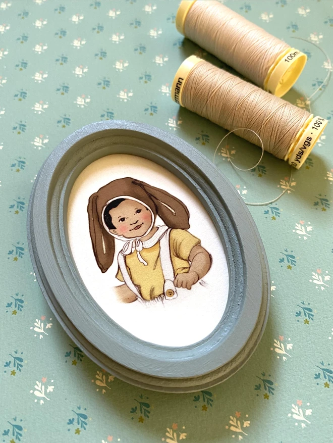 a photogrpah of a personalised drawing of a little asian boy wearing a easter bunny bonnet. framed in a duck egg blue oval wooden frame