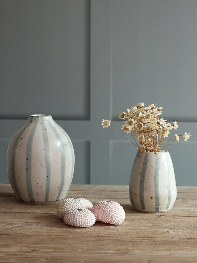 Handmade Ceramic Stoneware Vases in Blue and White Stipe in three different sizes: Small, medium and large sat on a wooden surface. The largest vase holds a bouquet of pink, yellow, red and white flowers. 