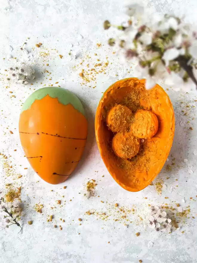 An orange carrot easter egg sitting on top of a blue background with a half open egg showing chocolate covered macarons embedded inside