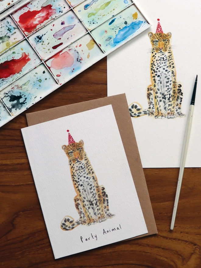 Desk Shot Of The Party Animal Leopard Greeting Card Accompanied By The Original Painted Illustration, Paint Brush And Paint Palette